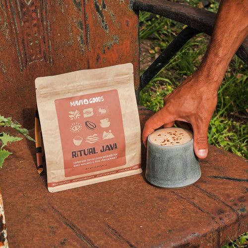 Hand reaching for a cup of coffee next to a package labeled 'Ritual Java' on a rustic wooden surface outdoors.

